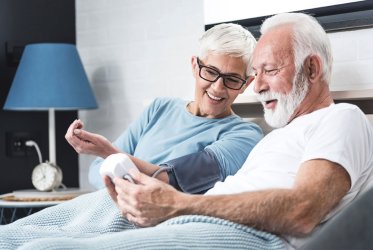 Ein älteres Paar liegt auf dem Bett. Der Mann misst den Blutdruck bei der Frau, die die Manschette an ihrem Oberarm platziert hat. Neben ihnen steht ein Tisch, auf dem eine Lampe und ein Wecker stehen.