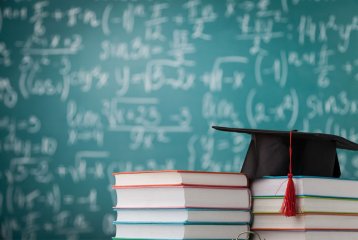 Ein Stapel mit Büchern, auf dem ein Mortarboard liegt. Im Hintergrund ist eine Tafel, auf der mit weißer Kreide etwas geschrieben steht.