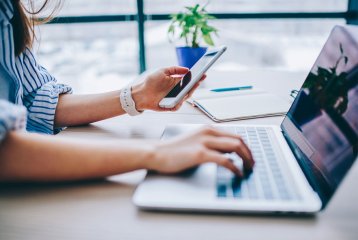 Eine Frau sitzt am Tisch, vor ihr ein Laptop. In der Hand hält sie ein Handy, auf das sie schaut. Neben ihr liegt ein Notizblock und eine grüne Pflanze