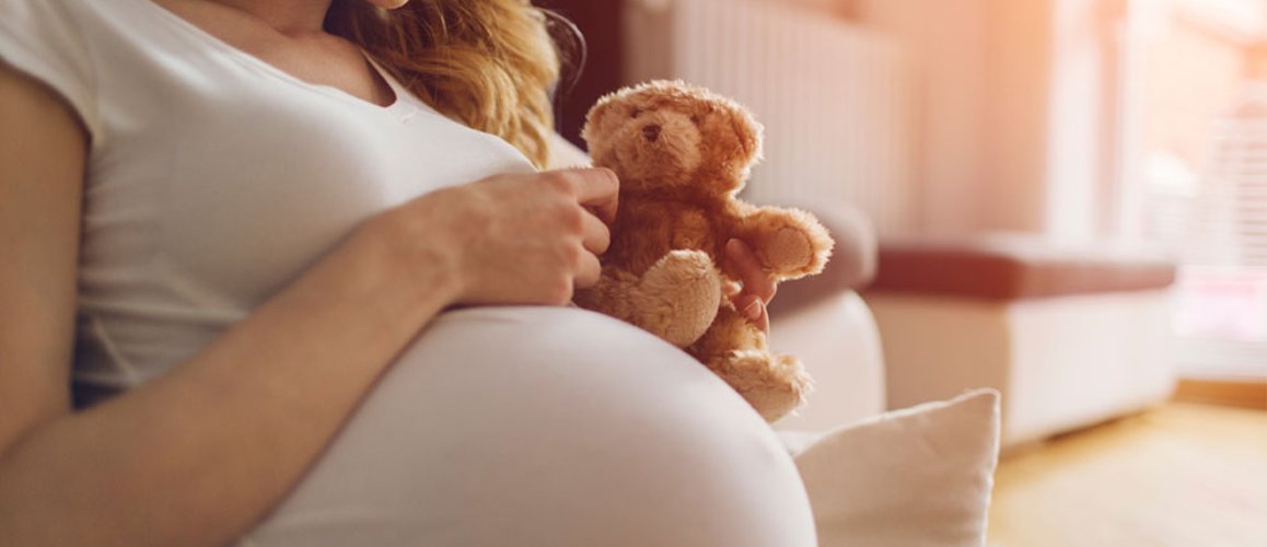 Eine schwangere Frau in einem weißen T-Shirt hält ein Kuscheltier in Form eines Bären in der Hand. Im Hintergrund ist der Abriss eines Wohnzimmers zu sehen.