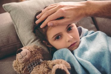 Ein Kind, das auf dem Sofa liegt, in einer blauen Decke, mit einer Hand an ihrem Kopf, misst Fieber. Sie sieht krank aus.