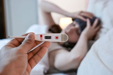 Eine Frau liegt krank im Bett, hält sich die Stirn mit einem Lappen, und ein Fieberthermometer wird in die Kamera gehalten. Eine Hand hält es.