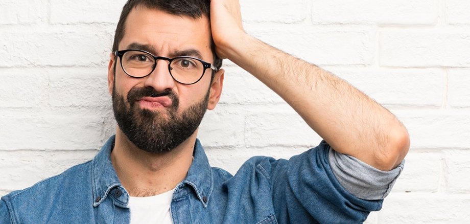 Ein Mann im weißen T-Shirt und blauen Hemd, der nachdenklich mit einer Hand an den Kopf fasst. Er steht vor einer weißen Wand und trägt eine Brille.