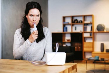 Eine Frau mit schwarzen Haaren sitzt an einem Tisch und inhalisiert mit einem Mundstück aus dem Inhalationsgerät, das auf dem hölzernen Tisch steht. Im Hintergrund ist ein Abriss eines Wohnzimmers zu erkennen.